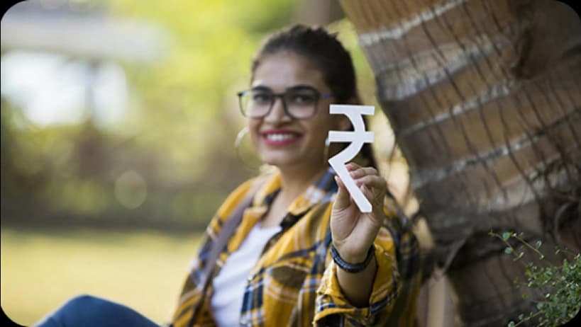 Smiling woman in glasses holds up a white Indian Rupee symbol outdoors.